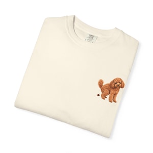 May include: A cream-coloured crewneck sweatshirt featuring a small, detailed graphic of a brown poodle dog on the lower right side. The dog is depicted in a standing position with a small brown pile behind it. The inner neck label reads Comfort Colors.