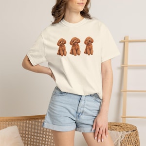 Puede incluir: Una camiseta de manga corta color crema con un estampado gráfico de tres caniches marrones sentados en fila. La prenda tiene cuello redondo y un corte relajado, combinada con pantalones cortos de mezclilla azul claro con dobladillo.