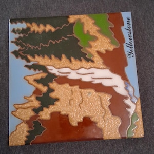 May include: A square ceramic tile featuring a stylized landscape illustration of Yellowstone. The design includes dark green pine trees, textured tan terrain, a white flowing river, and a light blue sky, with the word Yellowstone printed in black.