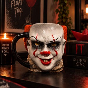 Puede incluir: Una taza de cerámica esculpida con la forma del rostro del payaso Pennywise, con cara blanca, nariz roja, marcas rojas en los labios, ojos amarillos y cabello naranja. La taza tiene un asa negra en el lado izquierdo y un cuello texturizado.
