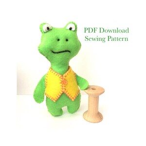 May include: A bright green felt frog doll wearing a yellow vest with orange stitching and a small orange button. Next to the frog is a light-colored wooden spool. The text PDF Download Sewing Pattern is visible in green on the right side.