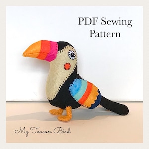 Peut inclure: Un jouet en peluche toucan en feutrine avec un corps noir, une poitrine beige et un bec multicolore aux sections orange et rose. L'aile présente des rayures bleues, orange et bleu foncé. Le texte indique PDF Sewing Pattern et My Toucan Bird.