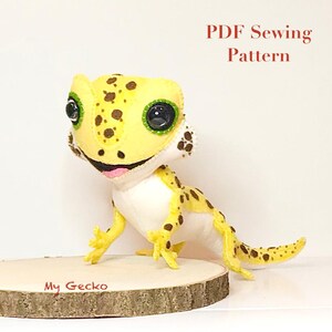 May include: A handcrafted felt gecko plush toy with yellow skin, brown spots, and large black eyes. The text 'PDF Sewing Pattern' and 'My Gecko' are visible. The gecko has a white underbelly, a small pink tongue, and is positioned on a round wooden base.