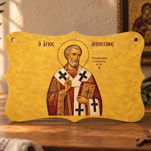 Könnte beinhalten: Eine rechteckige, goldfarbene Plakette mit geschwungenen Rändern, die eine religiöse Ikone des heiligen Augustinus zeigt. Die Figur trägt bischöfliche Gewänder mit schwarzen Kreuzen, hält ein Buch und zeigt griechischen Text.