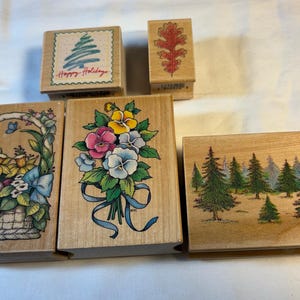May include: A collection of five wooden rubber stamps featuring various designs: a Christmas tree with the text Happy Holidays, an oak leaf, a basket of spring flowers, a bouquet of pansies with a blue ribbon, and a forest of pine trees.
