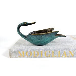 May include: A teal-colored swan-shaped decorative dish with a brass interior and gold accents. The swan is perched on a book with gold lettering. The dish has a textured finish and a curved neck.