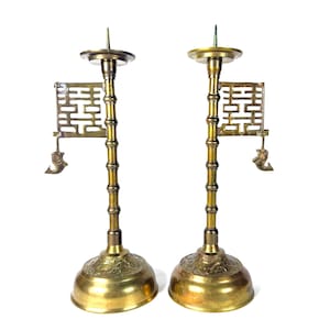 May include: Pair of antique brass candle holders. Each holder features a bamboo-style stem, a decorative base, and a small, ornate flag with a cutout design. The candle holders are a warm gold color and stand on a white background.