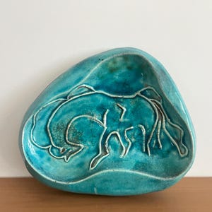 May include: A turquoise ceramic dish with a raised design of a horse. The dish is shaped like a teardrop and has a smooth, glazed finish.
