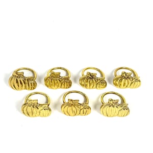 May include: A set of six gold-toned metal napkin rings, each featuring a sculpted pumpkin design with a circular band. The metallic finish is reflective, and the pumpkin motif includes detailed ridges and a small stem at the top of each piece.