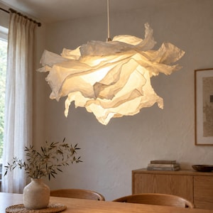 May include: A white pendant light fixture with a layered, ruffled, and translucent shade that emits a warm, soft glow. The structure is suspended from a single cord and features irregular, paper-like folds that create a voluminous, cloud-like shape.