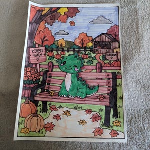 Op de afbeelding: Een ingekleurde tekening van een groene cartoon-dinosaurus die op een houten parkbank in een pompoenveld zit. De scène bevat oranje pompoenen, herfstbomen, een houten bord met de tekst KÜRBIS FARM en een kleine schuur onder een blauwe lucht.