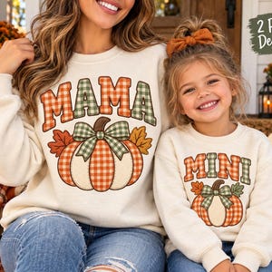 Peut inclure: Deux sweat-shirts à col rond couleur crème avec un motif de citrouille orné d'un nœud à carreaux et de feuilles d'automne. Le plus grand porte l'inscription MAMA en lettres à carreaux, et le plus petit porte l'inscription MINI.