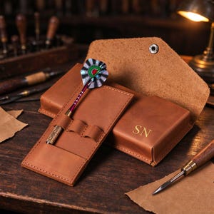 May include: A tan leather dart case with a flap closure and a metal snap button. The case features the embossed letters SN and includes a dedicated slot holding a single dart with a metallic purple shaft and a patterned flight in green, red, and silver.