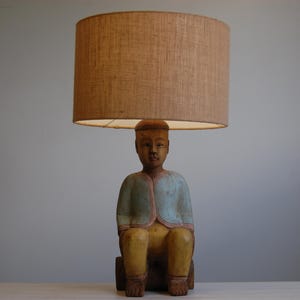 Może przedstawiać: Lampa stołowa z rzeźbioną drewnianą figurką osoby siedzącej na cylindrycznej podstawie. Figurka ma na sobie jasnoniebieską kurtkę i żółte spodnie. Na głowie figurki znajduje się duży, cylindryczny abażur z tkaniny w kolorze brązowym.