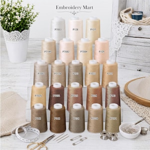 May include: A collection of twenty-one embroidery thread spools in various neutral beige, tan, and brown shades, each labeled with a product code. The spools are arranged on a tiered display stand, accompanied by sewing tools including scissors, needles, and a tape measure.