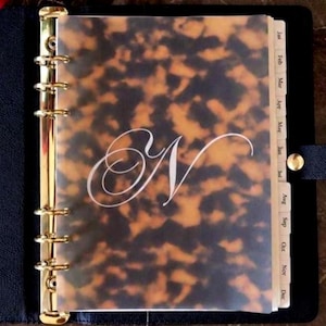May include: A black leather personal planner featuring a gold six-ring binder mechanism and a translucent tortoiseshell patterned cover. The cover displays a large white cursive letter N. The planner includes monthly tabbed dividers from Jan to Dec.
