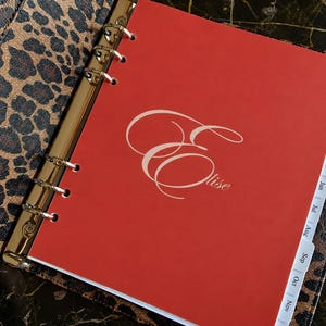 May include: A personal planner with a leopard print cover and a gold-toned six-ring metal binder mechanism. The interior features a bright red title page with the name Elise written in a white cursive script and white monthly tab dividers.