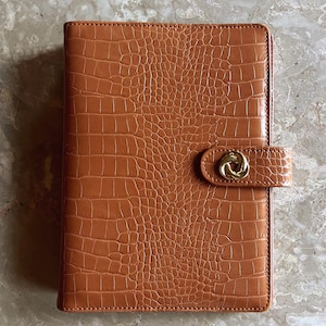 May include: A rectangular notebook with a tan, embossed crocodile-patterned cover. The notebook features a side strap closure secured by a circular, gold-toned metal knot-shaped button. The texture of the cover is uniform and glossy.