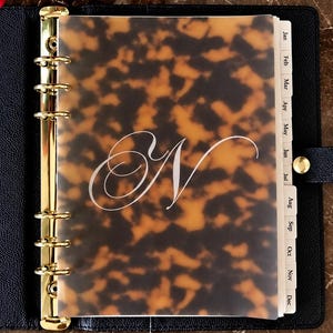 May include: A black leather planner with a gold six-ring binder mechanism and a translucent tortoiseshell patterned divider featuring a white cursive letter N. The planner includes monthly tabbed dividers labeled Jan through Dec.