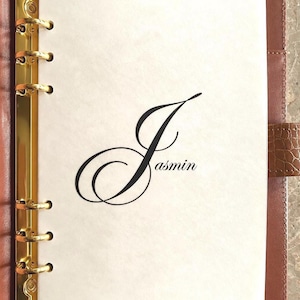 May include: A brown leather binder with a gold-colored six-ring metal mechanism. The interior features a white page with the name Jasmin printed in a large, elegant black script font, centered on the sheet. The binder has a textured cover.