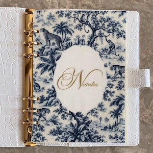 May include: A white faux leather planner with a crocodile texture and a gold six-ring binder mechanism. The translucent front page features a blue and white toile pattern with jungle animals and the name Natalia in gold script.
