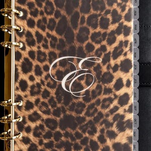 May include: A personal planner featuring a leopard print cover with a white cursive letter E in the center. The planner has gold-colored metal rings and gray monthly tab dividers labeled from January to December on the right side.