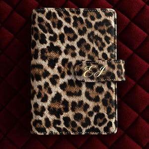 May include: A rectangular notebook cover featuring a brown and black leopard print pattern. The cover includes a side tab closure with the gold-colored cursive letters EJ embossed on the front. The texture appears to be short-haired.