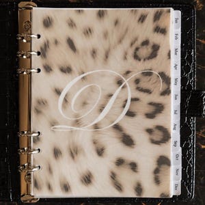 May include: A black crocodile-textured binder featuring a translucent divider with a beige and black leopard print pattern and a large white cursive letter D in the center, accompanied by monthly tabbed dividers from January to December.