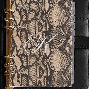 May include: A black leather planner featuring a gold six-ring binder mechanism and a translucent cover with a beige and black snakeskin pattern. The cover displays a large white cursive letter K and includes monthly tab dividers from Jan to Dec.