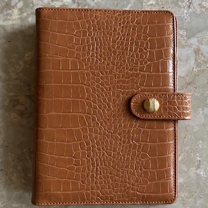 May include: A rectangular brown notebook with a textured crocodile skin pattern. The cover features a horizontal strap closure secured by a round, gold-toned metal button with a ribbed surface texture, centered on the right side.