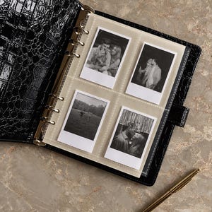 May include: A black binder with a glossy crocodile-embossed texture featuring a six-ring metal mechanism. Inside, a clear plastic page holds four black and white square photographs. A slim gold-toned metal pen rests beside the open binder.