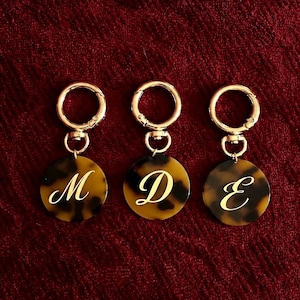May include: Three circular keychains featuring a brown and black tortoiseshell pattern. Each keychain has a gold-toned metal lobster clasp and a gold-toned letter engraved in the center, displaying the letters M, D, and E respectively.