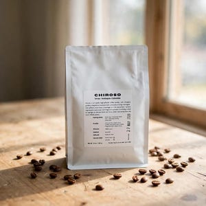 May include: A white coffee bag labelled Chiroso Urrao-Antioquia-Colombia with a net weight of 8 oz / 227 g and a harvest date of 27 May 2026. The bag is surrounded by scattered roasted coffee beans on a wooden surface, featuring detailed text.
