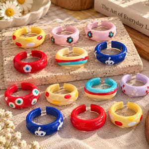 Puede incluir: Una colección de doce anillos abiertos de colores hechos de material liso. Los anillos presentan varios diseños, incluyendo colores sólidos, lunares, rayas horizontales y pequeños motivos florales en tonos rojo, azul, amarillo, rosa y morado.