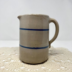 May include: A cylindrical stoneware pitcher with a light beige, speckled finish and two horizontal cobalt blue stripes encircling the body. The pitcher features a small pouring spout and a curved handle on the right side of the vessel.