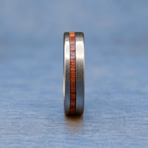 May include: A silver-toned metal ring featuring a central vertical inlay of reddish-brown wood with a visible grain pattern. The metal band has a brushed finish, and the wooden inlay is set flush within the center of the ring's outer surface.