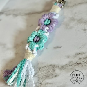 May include: A macrame keychain featuring two flower-shaped knots in lavender and mint green, accented with white cord. The design includes a cream-colored wrapped section near the silver metal clasp and a multi-colored tassel in purple, mint, and white.