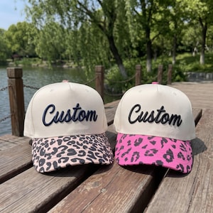 May include: Two baseball caps with off-white crowns and the word Custom embroidered in dark blue and black respectively. The left cap features a light pink and black leopard print brim, while the right cap has a bright pink and black leopard print brim.