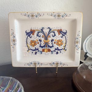 May include: A rectangular ceramic serving platter with an off-white base color. The center features a symmetrical floral and scrollwork design in shades of navy blue, mustard yellow, and burnt orange. The edges are accented with thin gold trim.