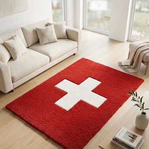 Puede incluir: Una alfombra rectangular que presenta un fondo rojo vibrante con un diseño de cruz blanca grande y centrado. La alfombra tiene una textura gruesa, afelpada y de pelo largo en toda su superficie, creando un aspecto suave y denso para el suelo.