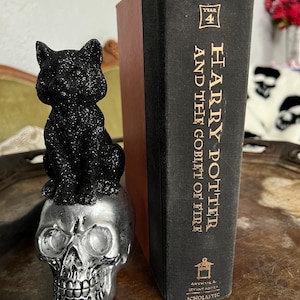 Może przedstawiać: Czarna figurka kota z brokatem siedząca na srebrnym czaszce-podstawce do książek. Podstawka do książek znajduje się obok czarnej książki w twardej oprawie z tytułem "Harry Potter i Czara Ognia" autorstwa J.K. Rowling.