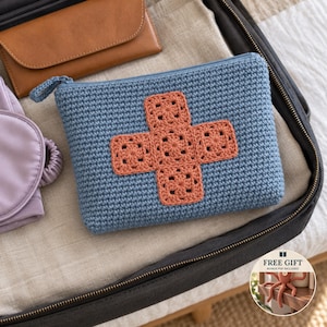 Op de afbeelding: Een blauw gehaakt tasje met ritssluiting en een kruisontwerp van vijf terracotta-kleurige granny squares op de voorzijde. Het tasje heeft een bijpassende blauwe gehaakte ritstrekker en een rechthoekige vorm met een textuur.