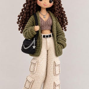Puede incluir: Una muñeca tejida a mano con largo cabello castaño rizado, gorra de béisbol beige con una M, top corto marrón, cárdigan verde oliva, pantalones cargo beige, cinturón negro y bolso bandolera negro con un dije de estrella plateada y cadena.