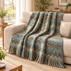 May include: A woven throw blanket featuring a multi-colored chevron pattern in shades of teal, navy blue, mustard yellow, and cream. The blanket has a textured knit construction and is finished with long, knotted fringe along the bottom edge.