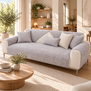 Może przedstawiać: Trzyosobowa sofa z pokrowcem z jasnoszarej, teksturowanej tkaniny oraz białymi panelami bocznymi i podłokietnikami. Sofa zawiera dwie kwadratowe szare poduszki dekoracyjne, jedną prostokątną szarą poduszkę lędźwiową i jedną kwadratową poduszkę w kolorze kremowym.
