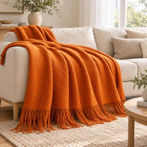 May include: A vibrant orange knit throw blanket featuring a textured bobble stitch pattern and long fringe detailing along the bottom edge. The blanket is draped over a light-colored sofa, showcasing its thick, woven construction and rich color.