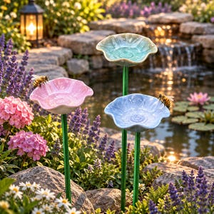 May include: Three ceramic flower-shaped garden stakes in pastel pink, light blue, and pale green, each mounted on a thin green metal rod. Two honeybees are perched on the edges of the pink and blue flower basins, which are designed to hold water.