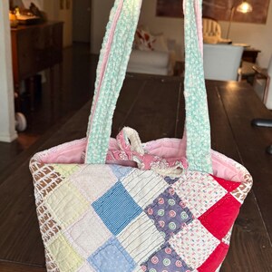 Puede incluir: Un bolso de mano acolchado hecho de varios cuadrados de tela en tonos rojo, azul, amarillo y blanco. El bolso cuenta con dos asas largas con estampado floral azul claro y un forro interior rosa con un pequeño cierre de lazo de tela.