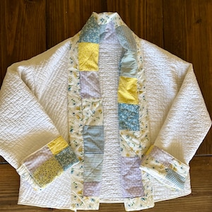 May include: A quilted jacket featuring a solid white main body with a grid-stitched texture. The front opening and sleeve cuffs are accented with a patchwork border of floral, gingham, and solid yellow, blue, and lavender fabric squares.