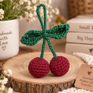 Puede incluir: Un par de cerezas de ganchillo de color rojo oscuro con dos hojas verdes de ganchillo y un tallo en forma de bucle verde para colgar. Se adjunta una pequeña etiqueta de papel rectangular marrón con un icono de corazón y el texto Crochet Makes Life Sweeter.
